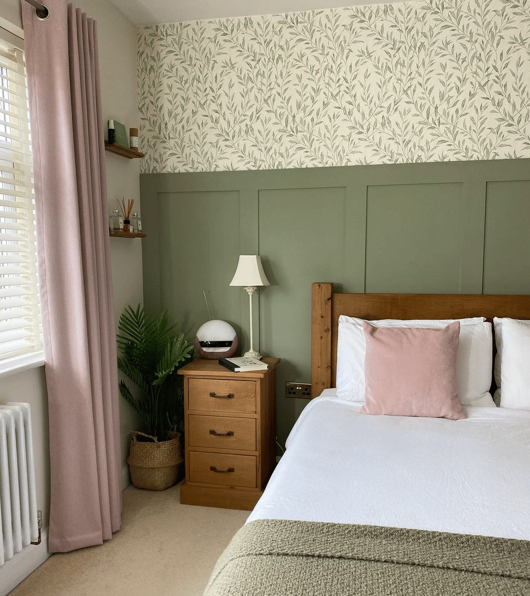 olive green half wall paneling with green foliage style wallpaper above it, there are pink curtains and a wooden bed frame in front of the paneling