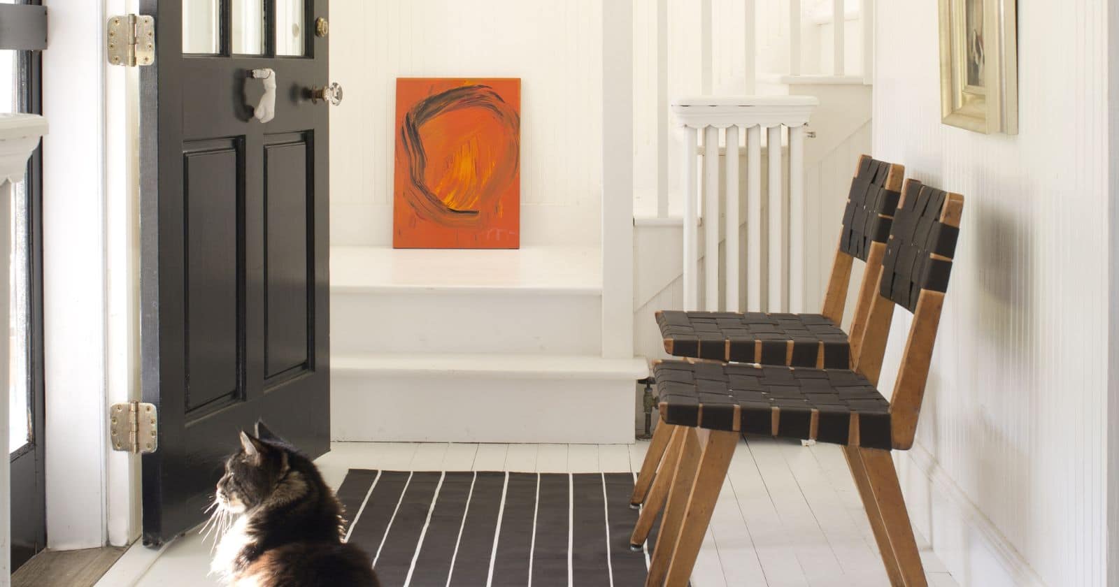 Bright white hallway with black front door, striped rug, wooden chairs, rustic light fixture, and a cat in sunlight.