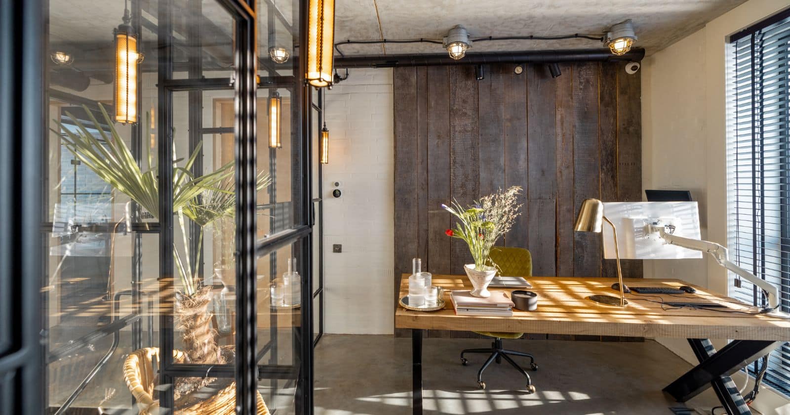 Industrial-style home office with glass partitions, wood accents, warm lighting, and a large desk under a concrete ceiling.