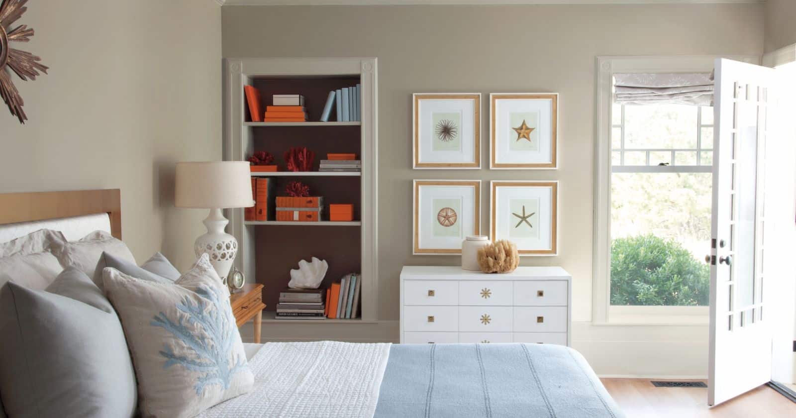 A bedroom with light walls, a large bed with blue and white bedding, a white dresser, and framed nautical prints.