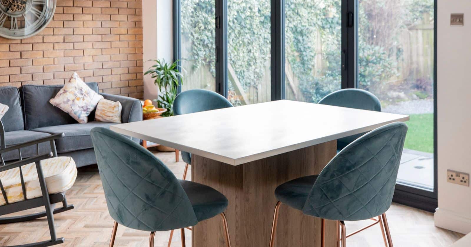 Close-up of a minimalist kitchen-diner with teal quilted bar stools, a pale wood island, and a view of the garden through black-framed glass doors.