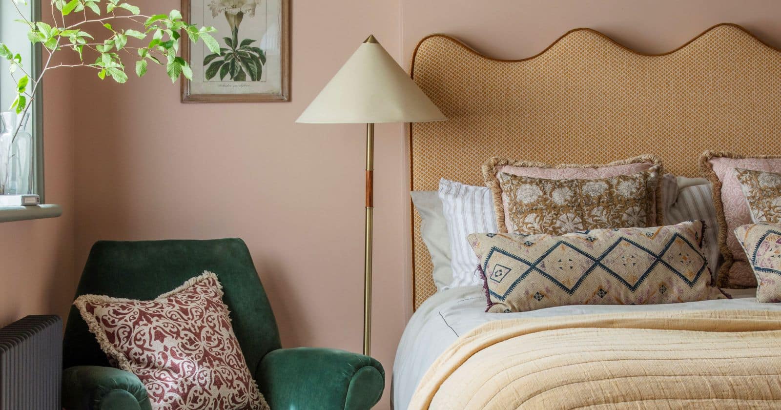 Cozy bedroom with blush pink walls and a scalloped mustard upholstered headboard. The bed is layered with embroidered cushions and a quilted blanket. A green velvet armchair with a patterned throw pillow sits beside a floor lamp and framed botanical print.