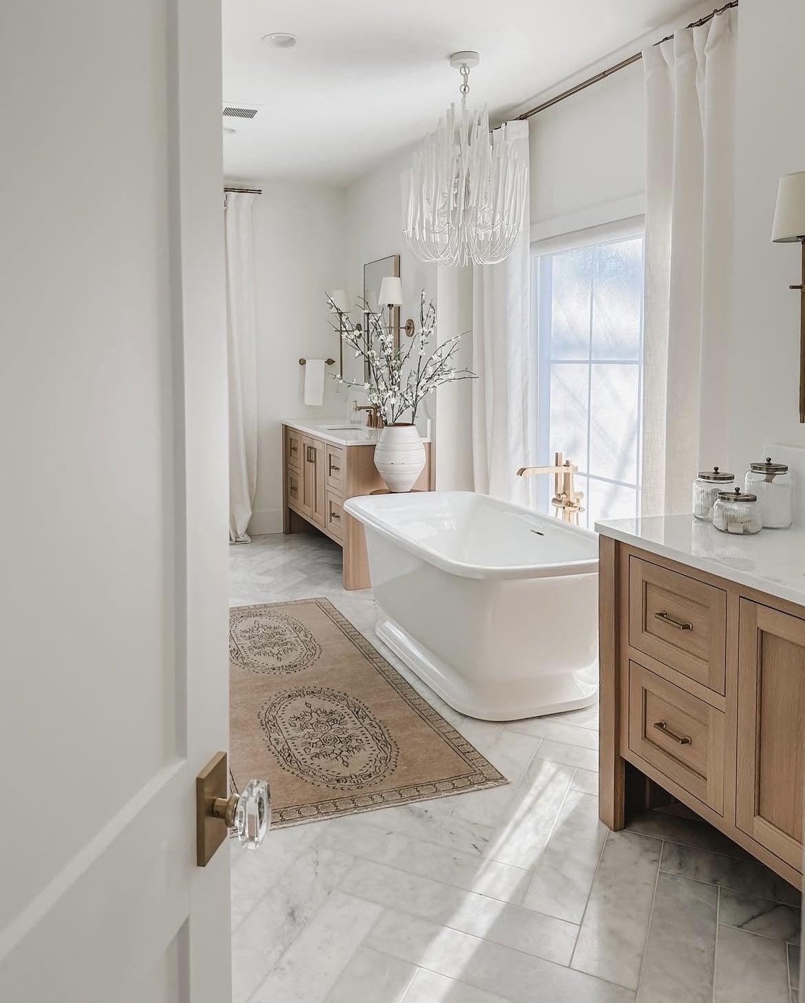 Elegant bathroom with a freestanding bathtub, marble floor tiles, natural wood cabinetry, chandelier, and large window with sheer curtains.