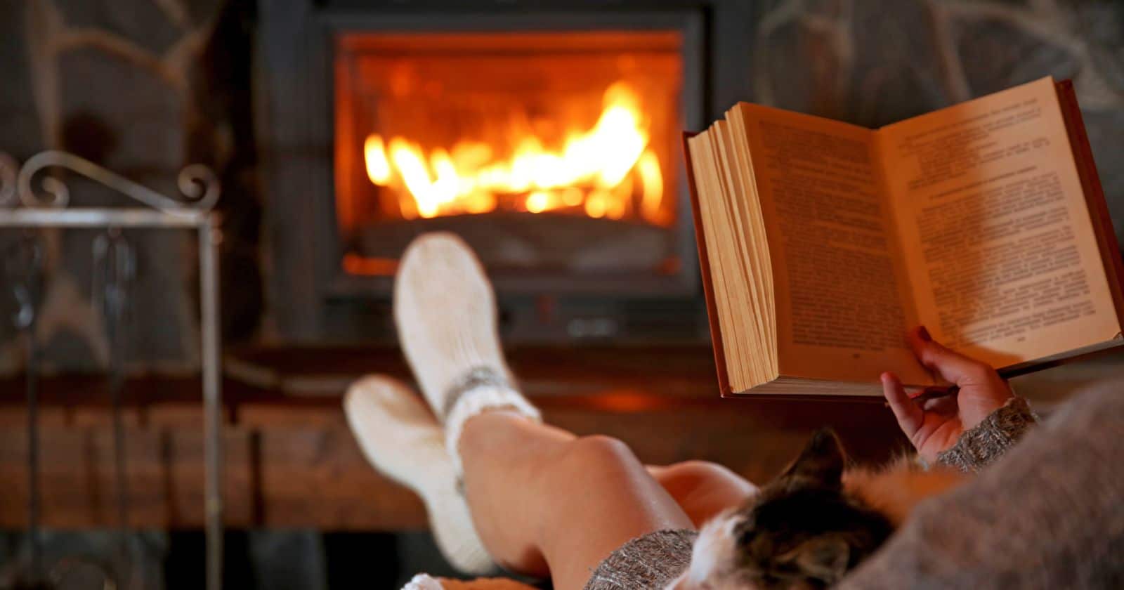 A person's feet in cozy socks warming up by a wood-burning stove while reading a book.
