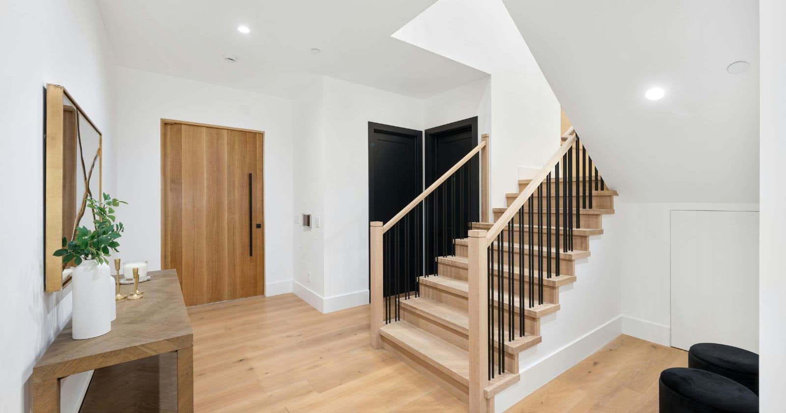 A spacious entryway featuring a light wood front door, two black doors, and a wood staircase with black balusters.